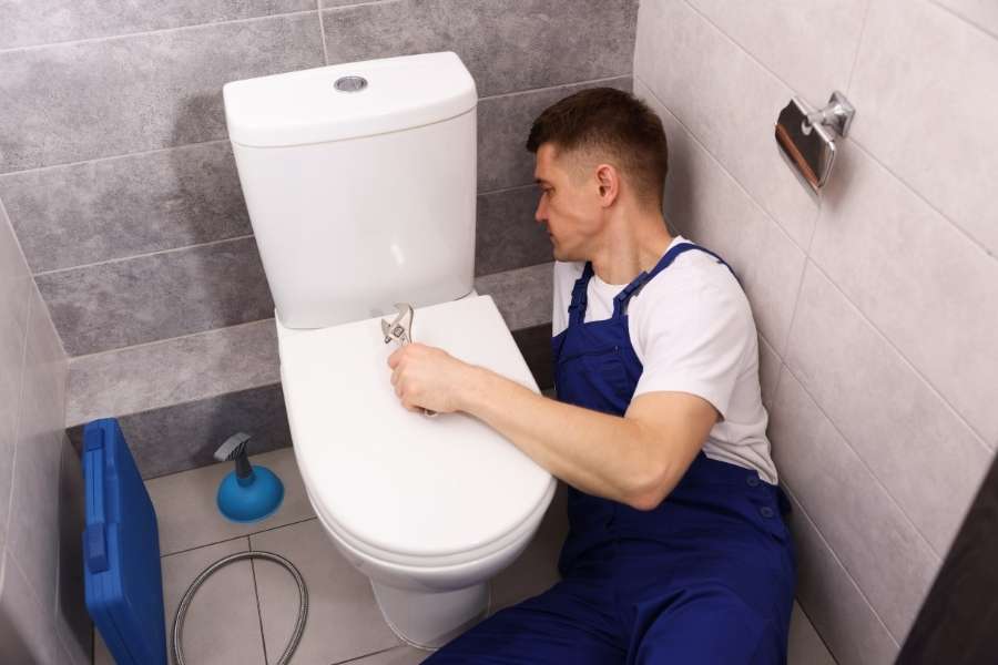 A professional plumber in blue overalls uses a wrench to tighten hardware on a toilet in a modern tiled restroom to fix a running toilet.
